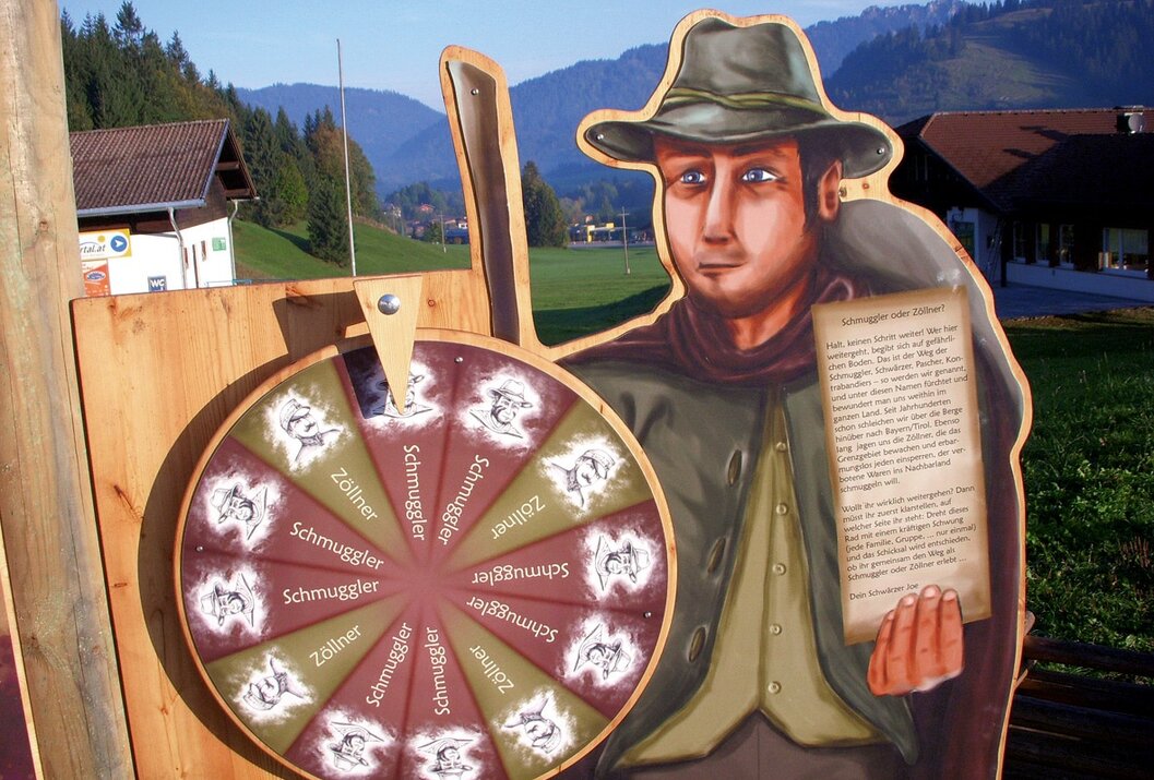 Holzfigur mit Hut und Umhang hält ein Glücksrad und ein Schriftstück vor Bergkulisse
