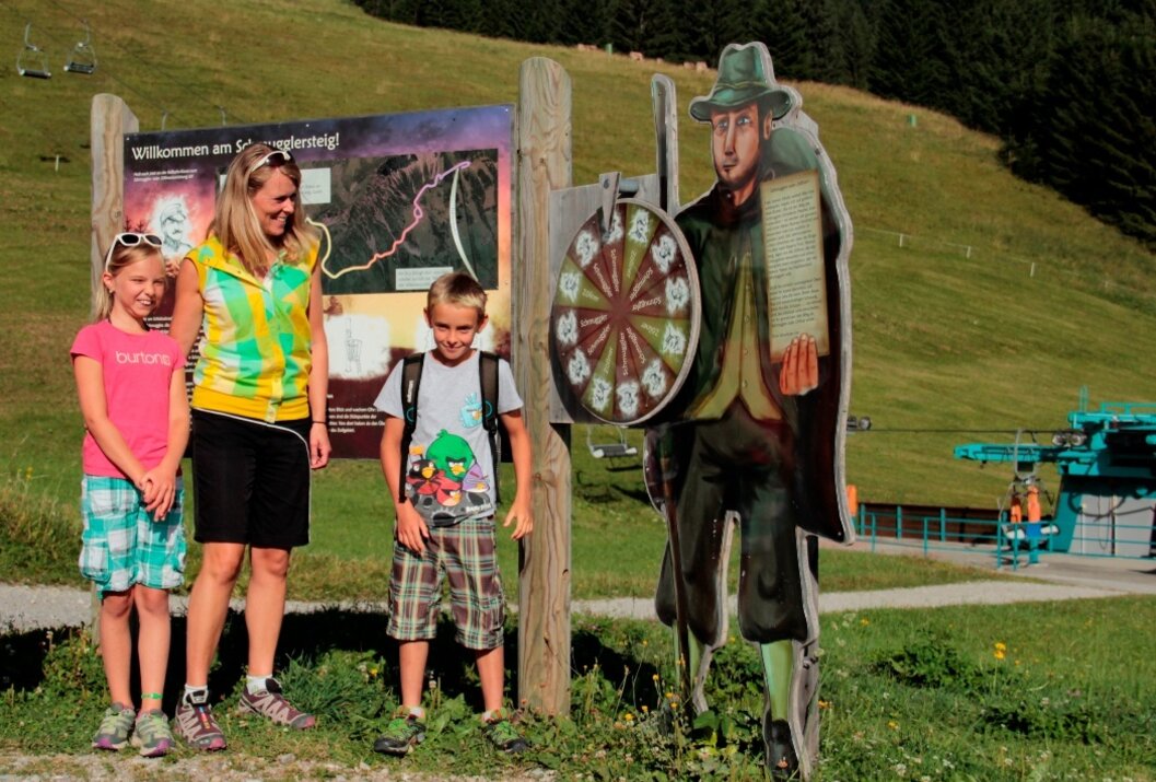 Familie posiert neben Holzfigur mit Dartscheibe vor Berglandschaft