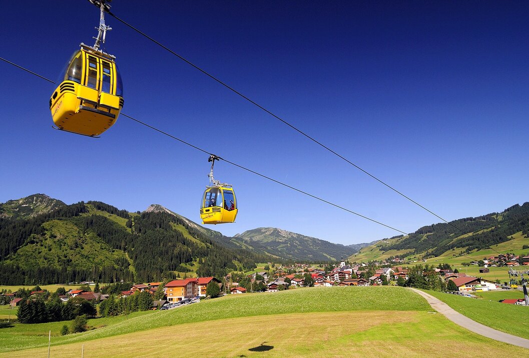 Gelbe Gondeln schweben über grüne Hügel und ein Dorf vor Bergkulisse unter blauem Himmel