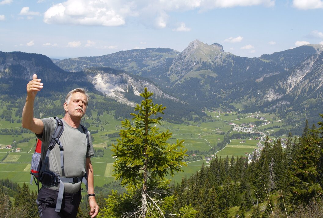 Wanderer vor Bergpanorama mit grünen Wiesen und bewaldeten Hängen unter blauem Himmel mit Wolken