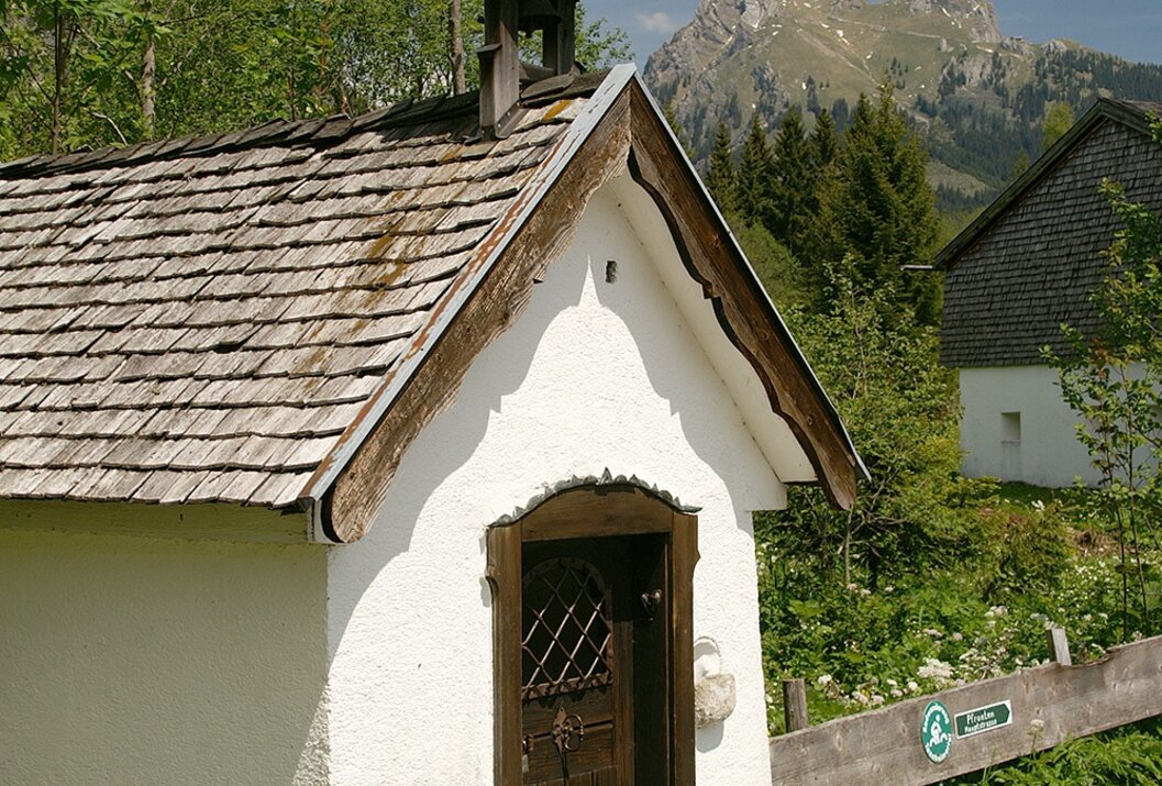 Kleine weiße Kapelle mit Holztür vor Bergkulisse und bewaldeter Umgebung