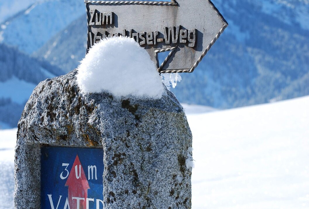 Wegweiser im Schnee mit Aufschrift "Zum Vater Unser Weg" vor Bergkulisse