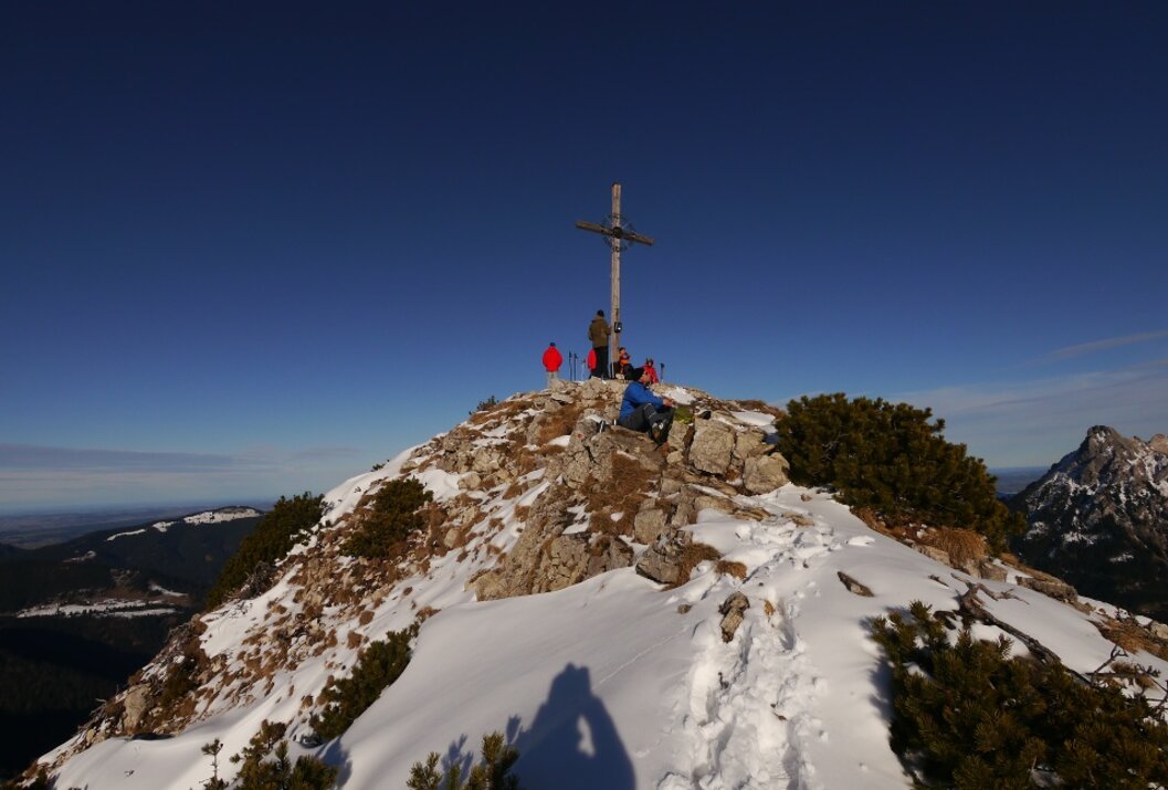Gipfelkreuz auf schneebedecktem Berg, Menschen stehen und sitzen daneben, blauer Himmel