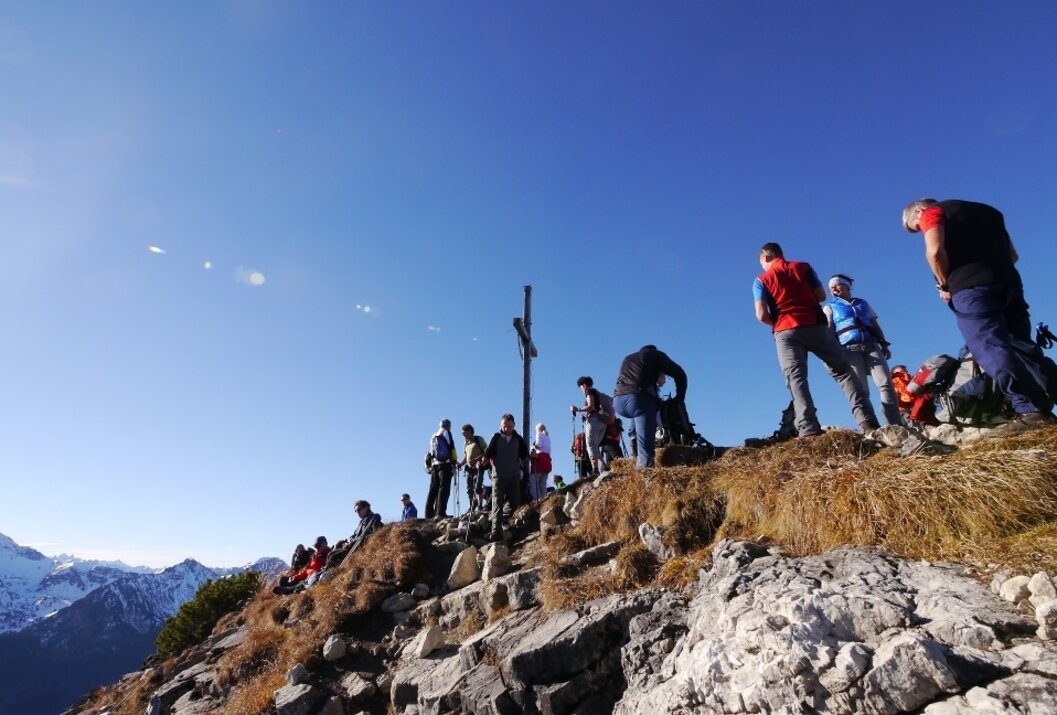 Menschengruppe auf einem felsigen Berggipfel mit Gipfelkreuz unter blauem Himmel
