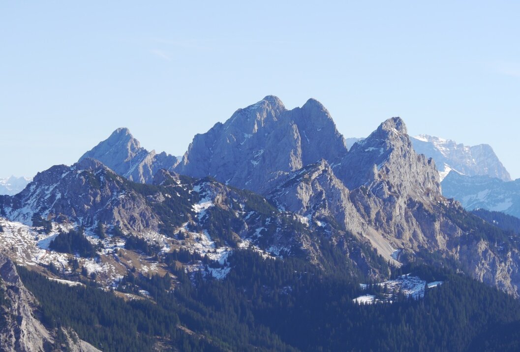 Bergpanorama mit schneebedeckten Gipfeln und bewaldeten Hängen unter blauem Himmel