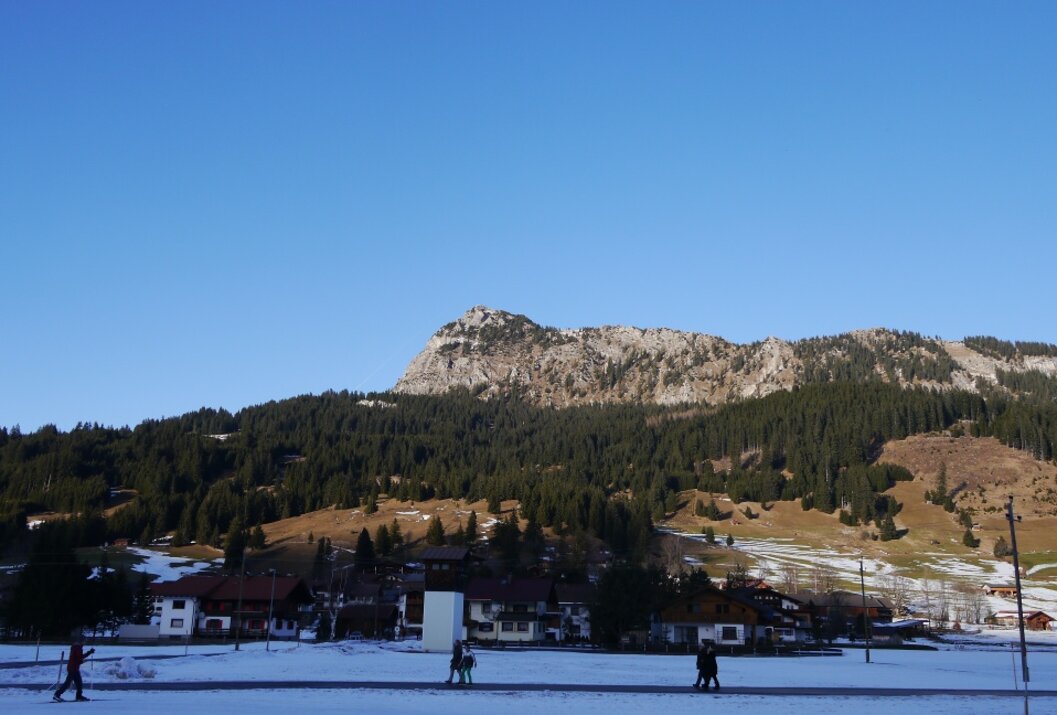 Berglandschaft mit schneebedeckten Hängen, dunklem Wald und Häusern im Vordergrund unter blauem Himmel