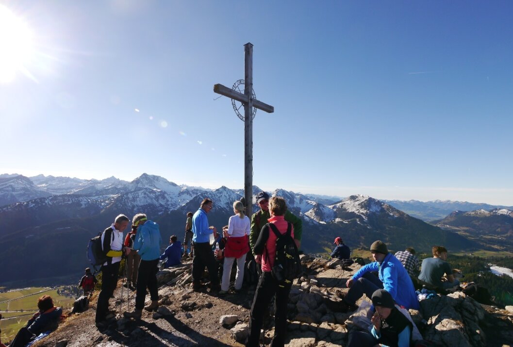 Wanderer stehen um ein Gipfelkreuz mit Bergpanorama im Hintergrund