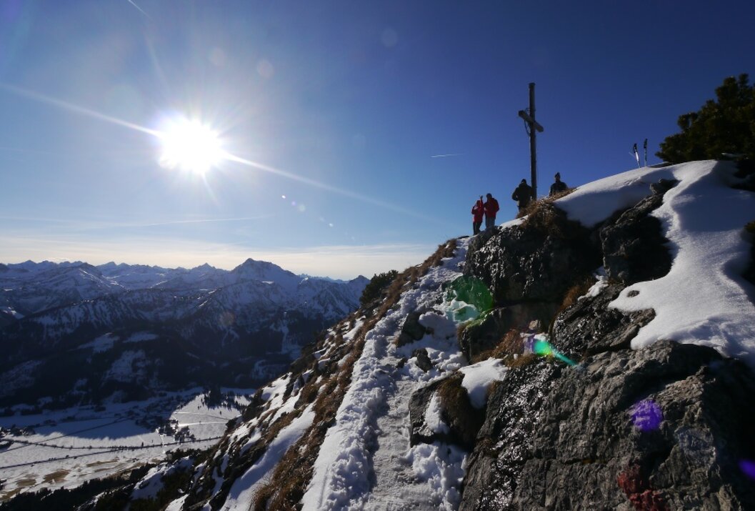 Verschneiter Berggipfel mit Gipfelkreuz, Wanderern und strahlender Sonne am blauen Himmel
