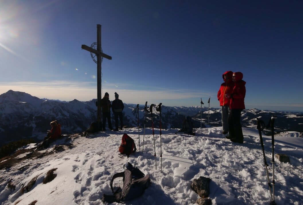 Gipfelkreuz auf schneebedecktem Berg, Wanderer in roter Jacke, Wanderstöcke im Schnee