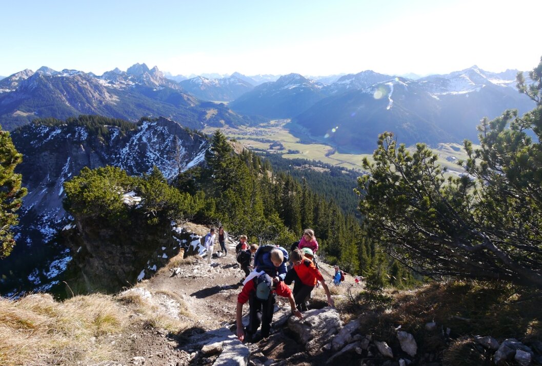 Wandergruppe auf Bergpfad mit Blick auf bewaldetes Tal und schneebedeckte Berggipfel im Hintergrund
