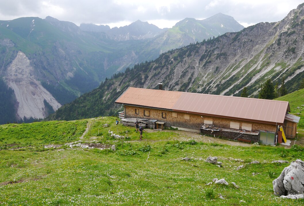 Hütte in Berglandschaft mit grüner Wiese im Vordergrund und bewaldeten Bergen im Hintergrund