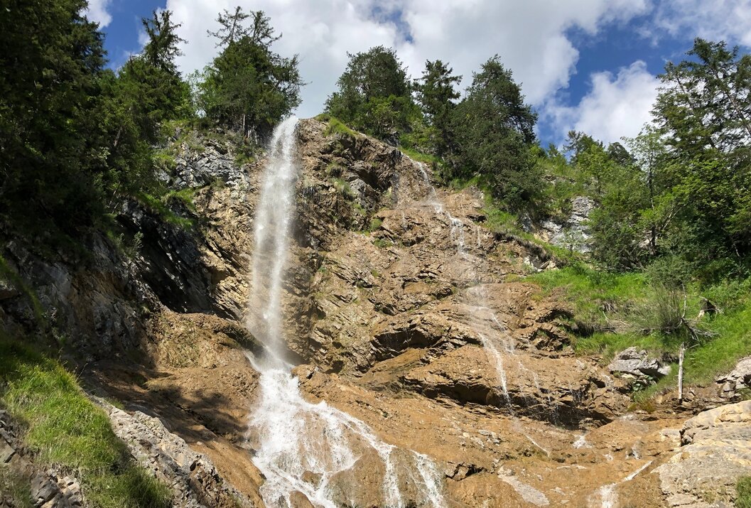 Wasserfall stürzt über Felsen, umgeben von Bäumen und blauem Himmel mit Wolken