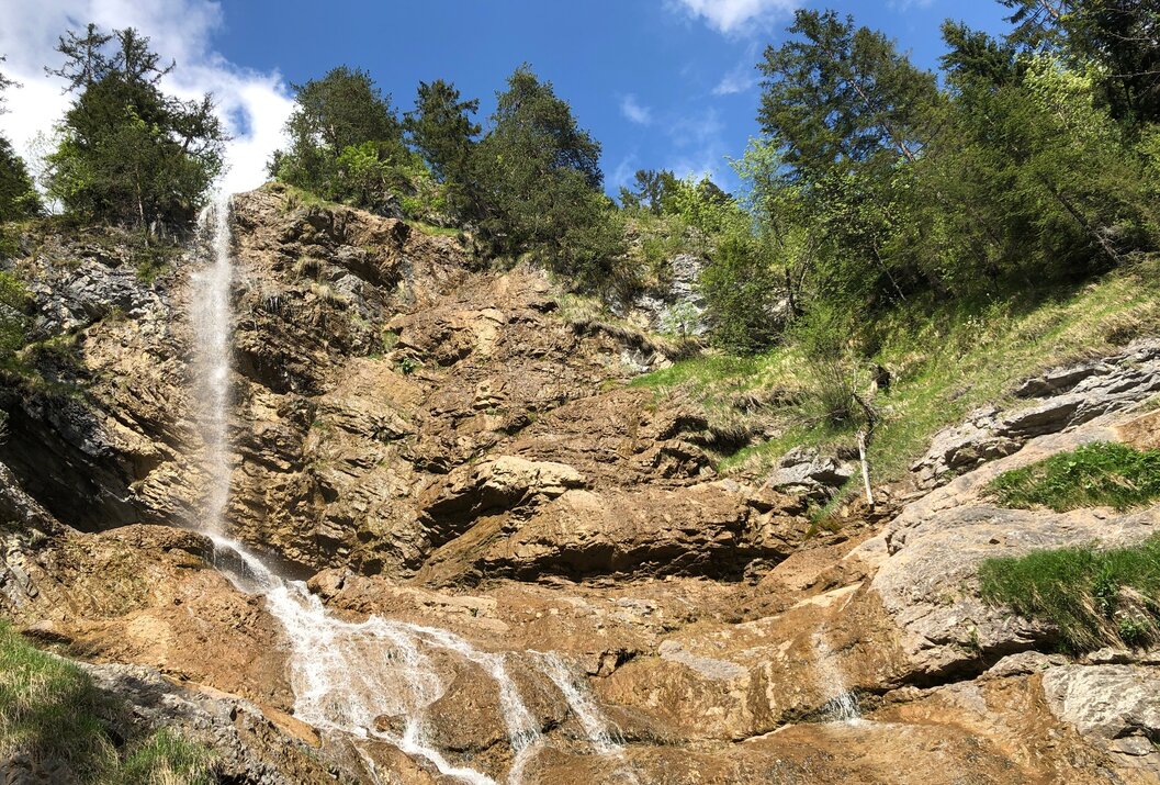 Wasserfall fließt über Felsen, umgeben von Bäumen und Grün unter blauem Himmel