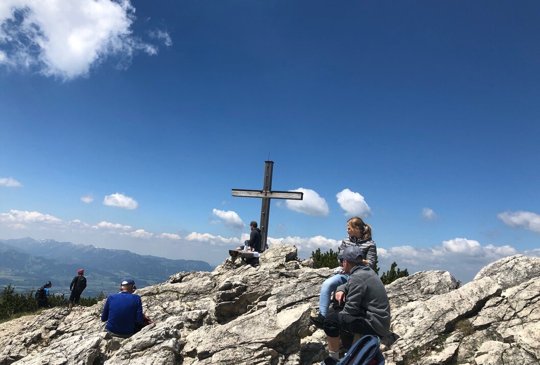 Menschen sitzen und stehen auf einem felsigen Berggipfel mit Gipfelkreuz unter blauem Himmel