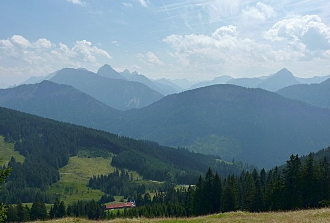Berglandschaft mit bewaldeten Hügeln und einem kleinen Haus im Tal unter blauem Himmel