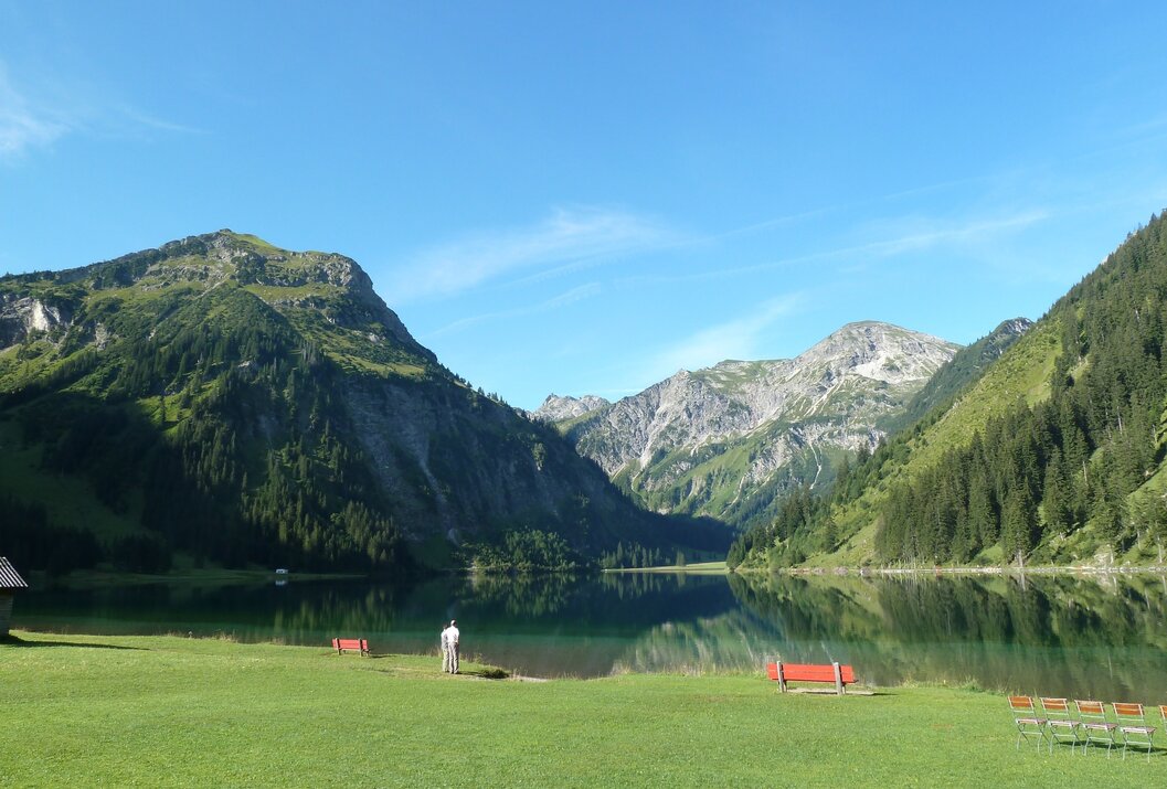 Bergsee mit grüner Wiese, Bergen im Hintergrund und blauem Himmel