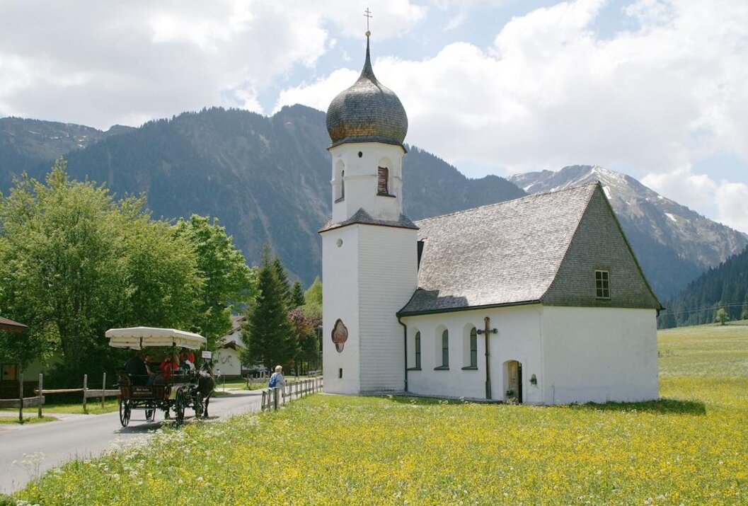 Weiße Kirche mit Zwiebelturm vor Bergkulisse, im Vordergrund eine Pferdekutsche