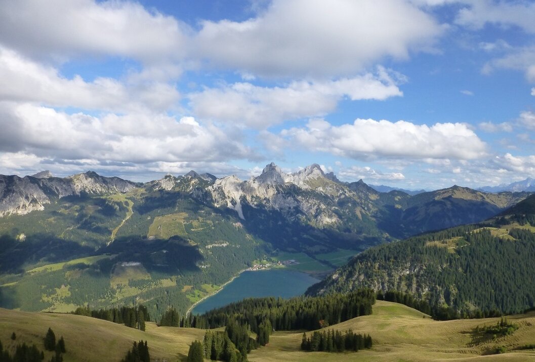 Berglandschaft mit See, Wiesen und bewaldeten Hängen unter blauem Himmel mit Wolken