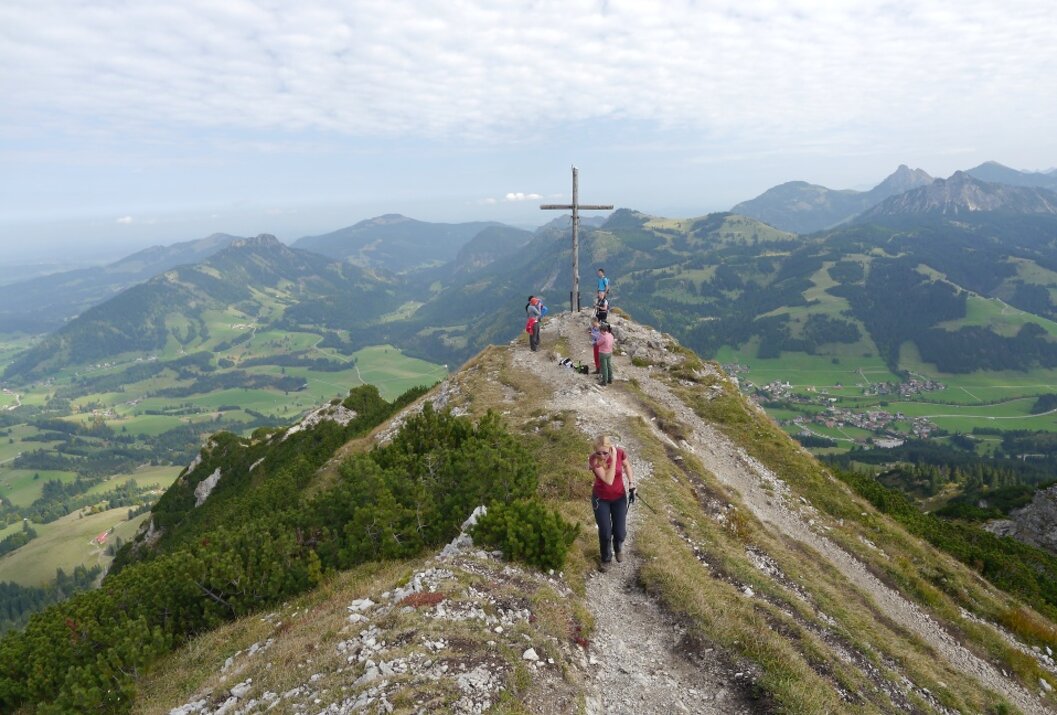 Wanderer auf Bergpfad zum Gipfelkreuz mit Panoramablick auf grüne Hügel und Tal