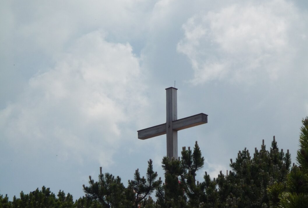 Hölzernes Gipfelkreuz über Baumwipfeln vor bewölktem Himmel