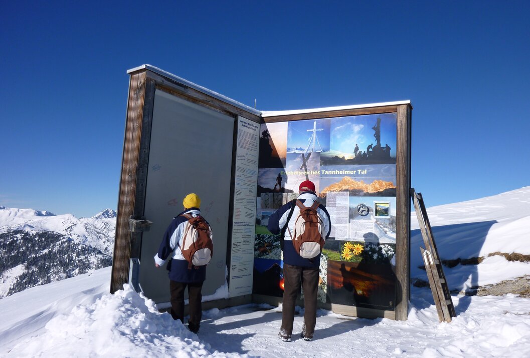 Zwei Wanderer betrachten Infotafel auf schneebedecktem Berggipfel unter blauem Himmel