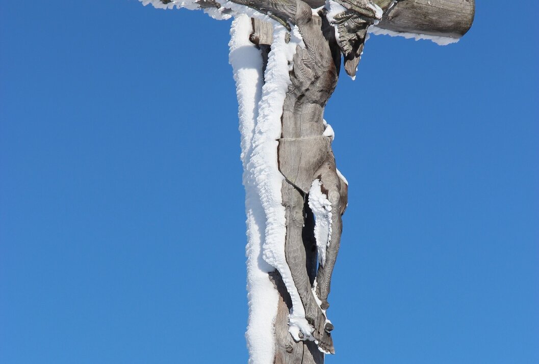 Holzkreuz mit Jesusfigur, teilweise mit Schnee bedeckt, vor blauem Himmel