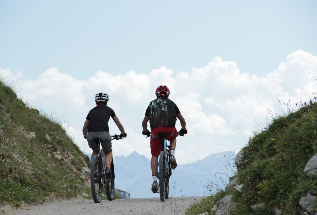 Zwei Mountainbiker fahren bergauf auf einem Schotterweg vor Bergkulisse
