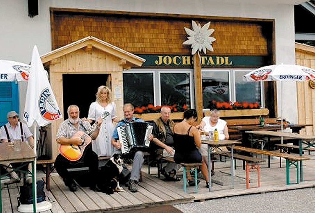 Gruppe vor dem Jochstadl mit Musikern, Hund und Gästen an Tischen