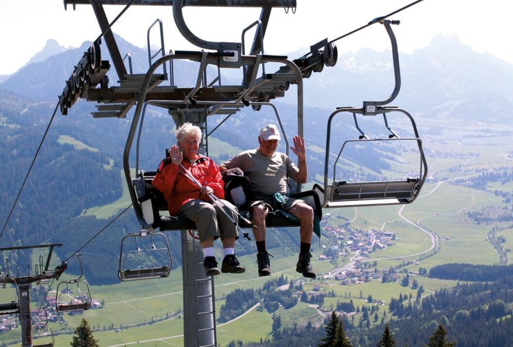 Älteres Paar winkt in einem Sessellift vor Bergpanorama