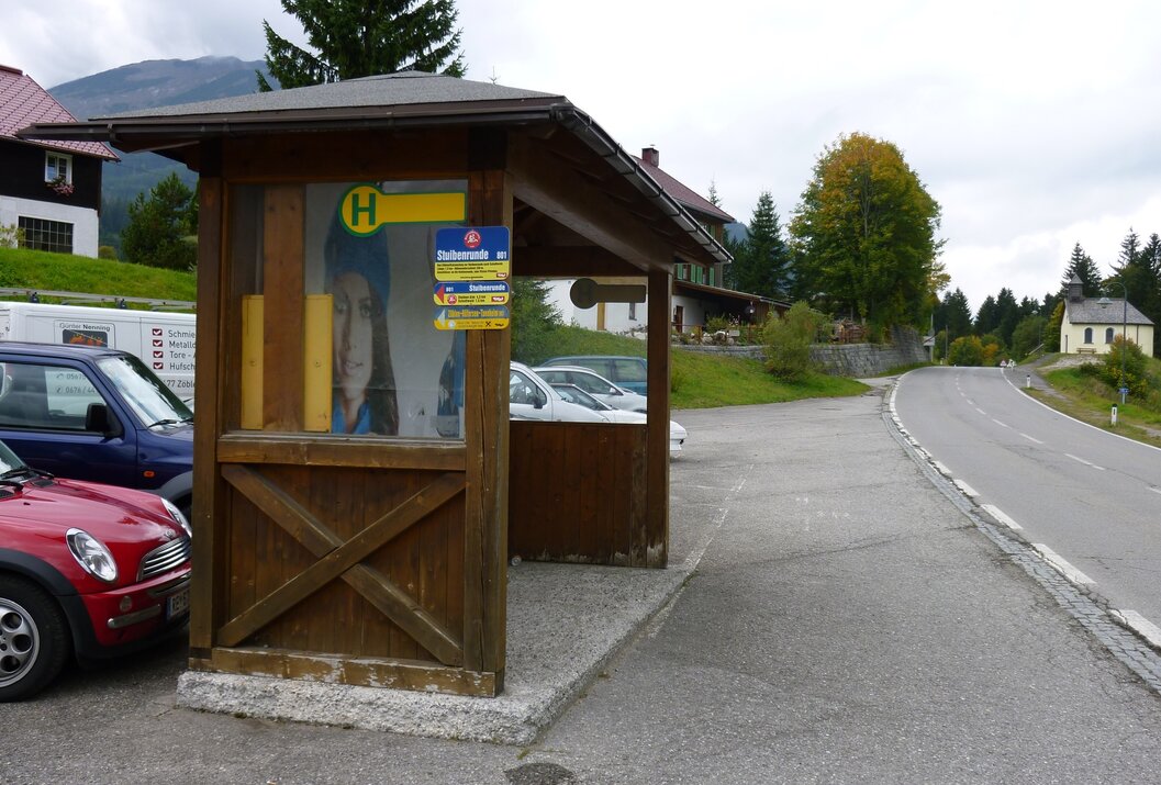 Hölzerne Bushaltestelle mit Fahrplan an einer Straße vor Häusern und Bergen