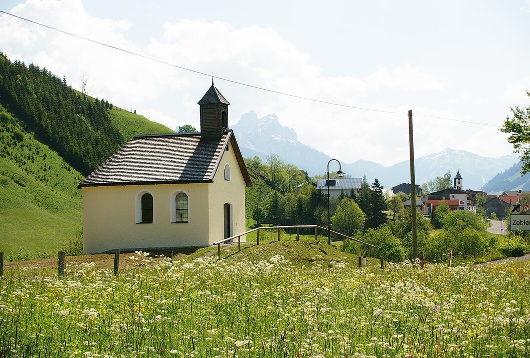 Kleine Kirche auf Hügel mit Blumenwiese, im Hintergrund Berge und Häuser