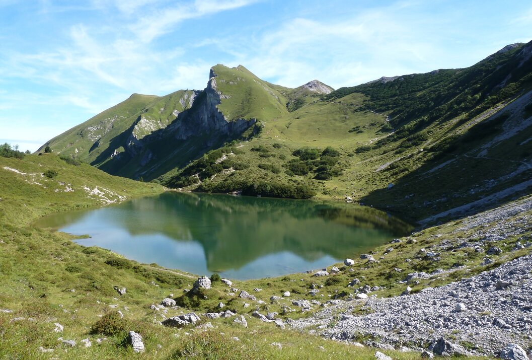 Bergsee umgeben von grünen Hügeln und Felsen unter blauem Himmel