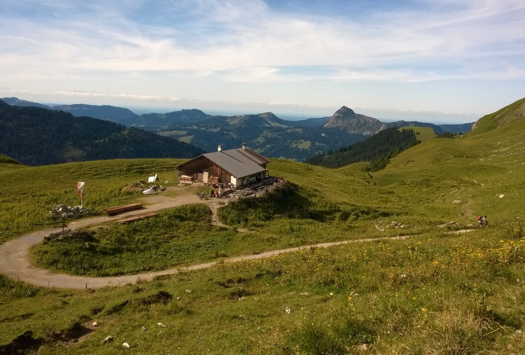 Hütte auf grüner Bergwiese vor Bergpanorama unter blauem Himmel