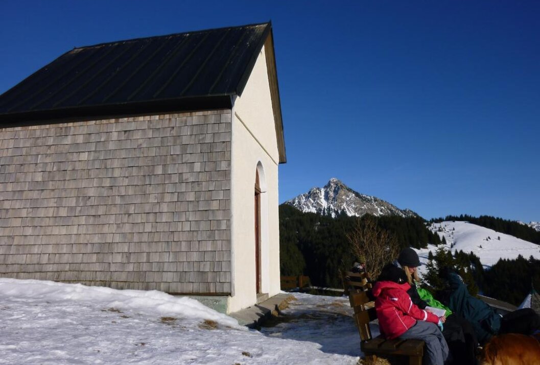 Kleine Kapelle im Schnee, davor Bank mit Personen, im Hintergrund Berg und blauer Himmel