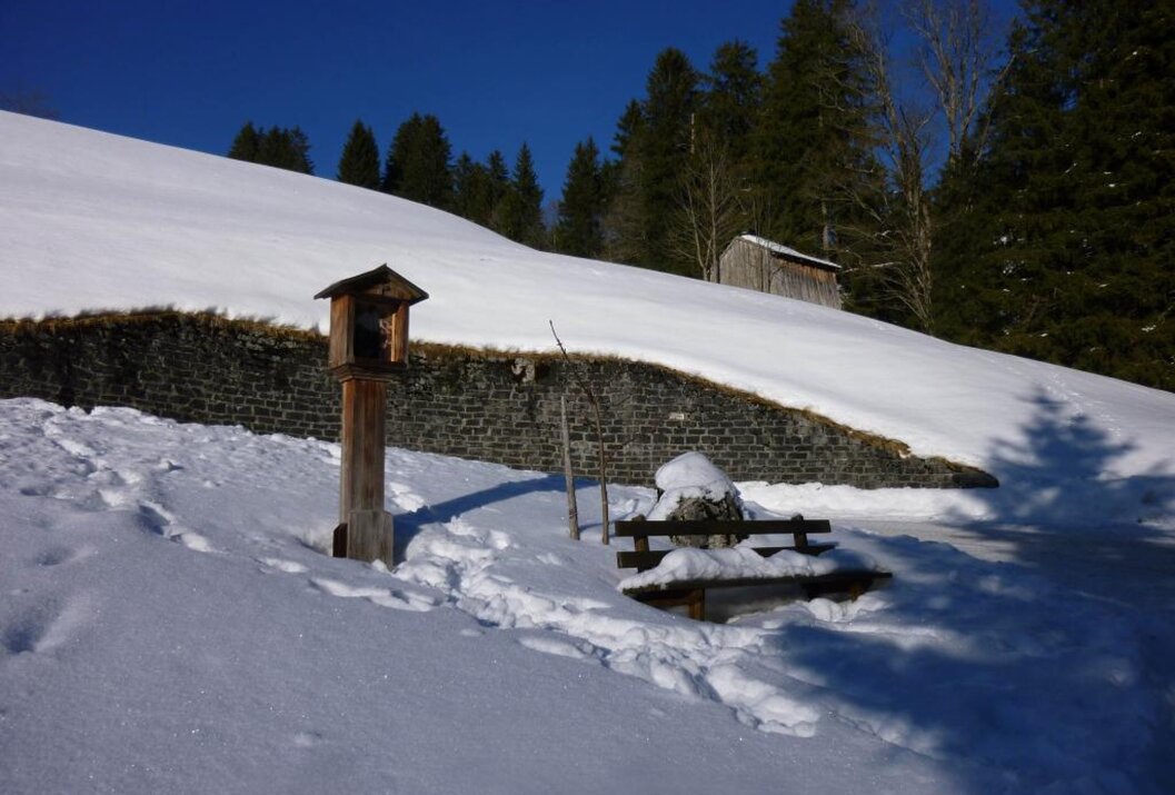 Schneebedeckte Landschaft mit Holzhäuschen, Bank und Wald im Hintergrund unter blauem Himmel