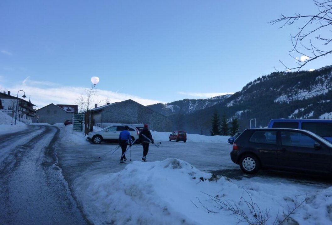 Zwei Skifahrer auf schneebedecktem Parkplatz vor Häusern und Bergen unter blauem Himmel