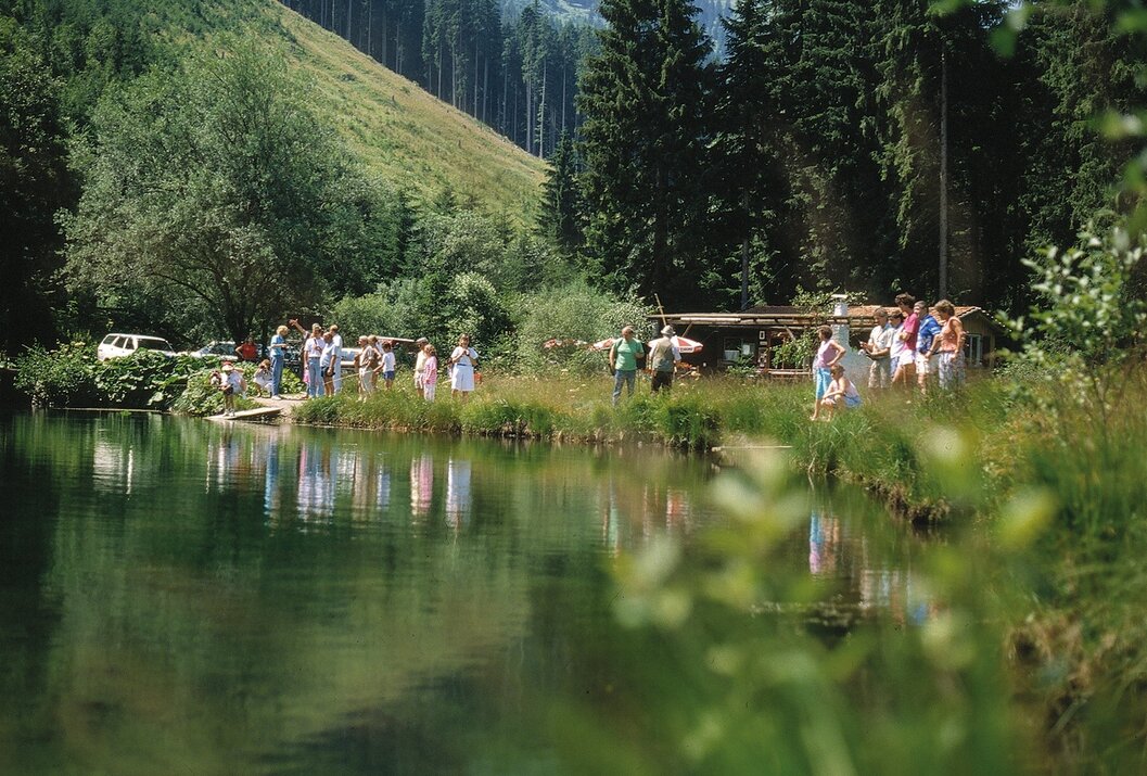 Menschengruppe am Ufer eines Bergsees mit Bäumen und Bergen im Hintergrund
