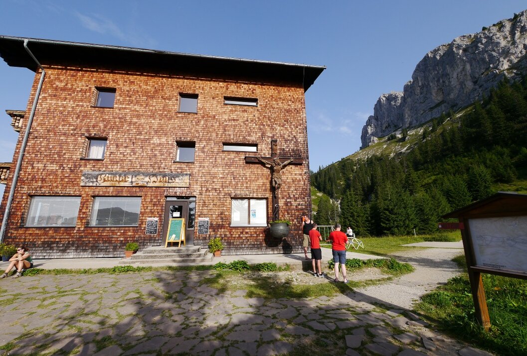Hütte mit Holzwand, Kreuz, Wanderern und Berglandschaft