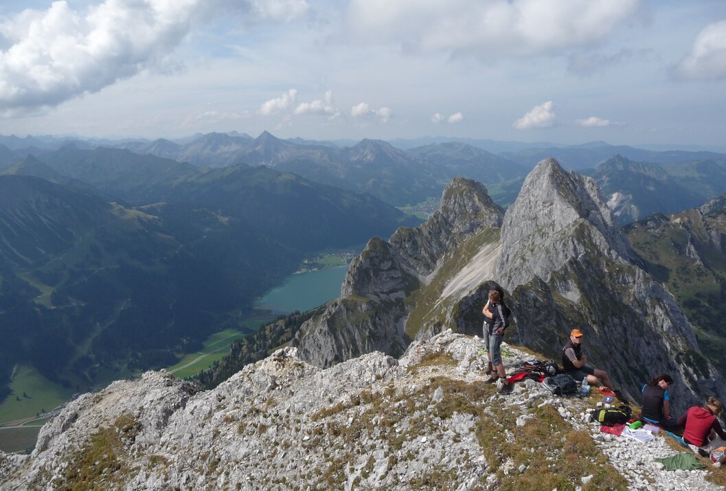 Berglandschaft mit zwei Wanderern auf einem felsigen Grat und einem See im Tal