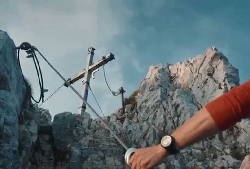 Gipfelkreuz auf felsigem Berg, Arm mit Uhr im Vordergrund, blauer Himmel