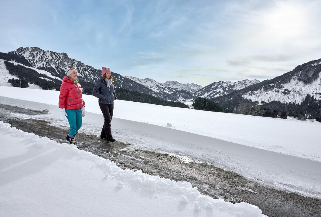 Zwei Frauen wandern auf einem schneebedeckten Weg vor Bergpanorama