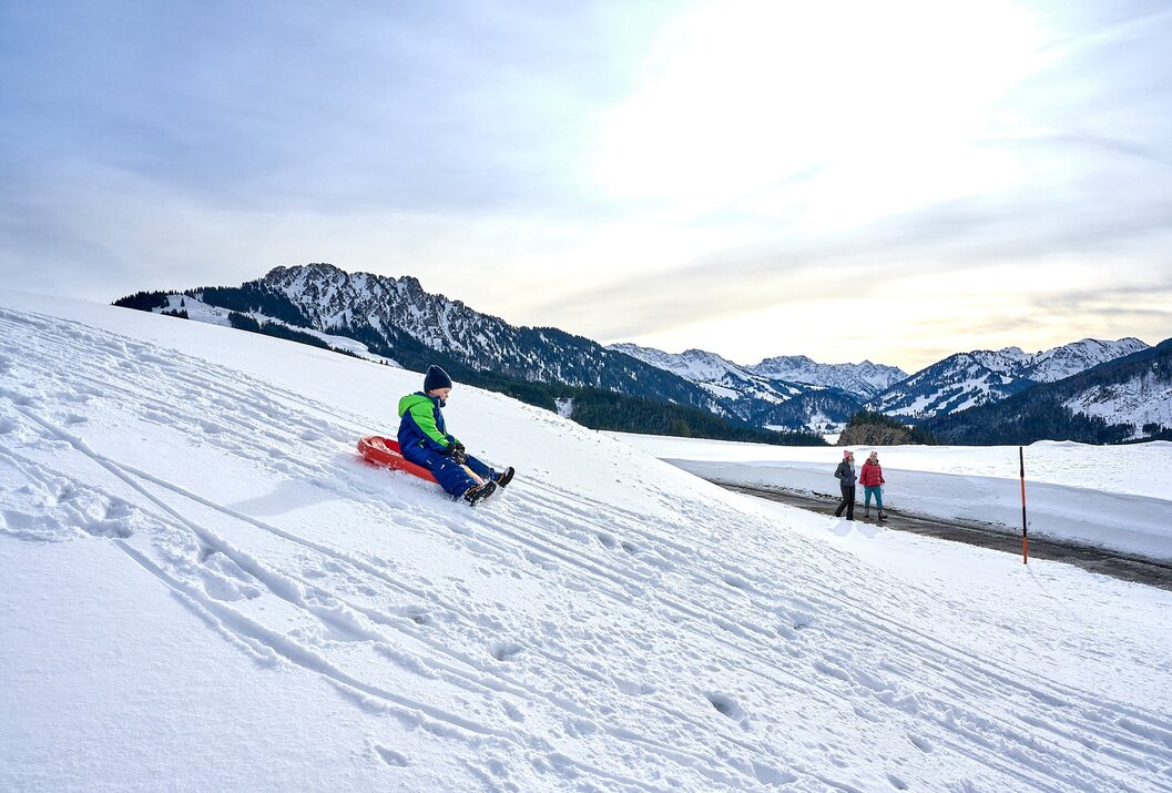 Kind fährt mit Schlitten einen schneebedeckten Hang hinunter, im Hintergrund Berge und zwei Personen