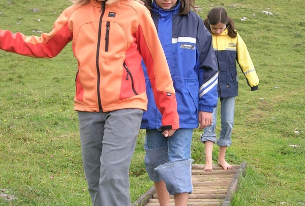 Drei barfüßige Mädchen gehen auf einem Holzsteg über eine grüne Wiese