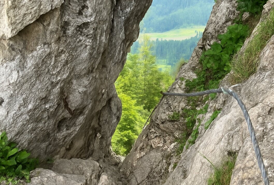 Felsiger Pfad mit Drahtseilversicherung führt durch eine Felsspalte mit Blick auf grüne Landschaft