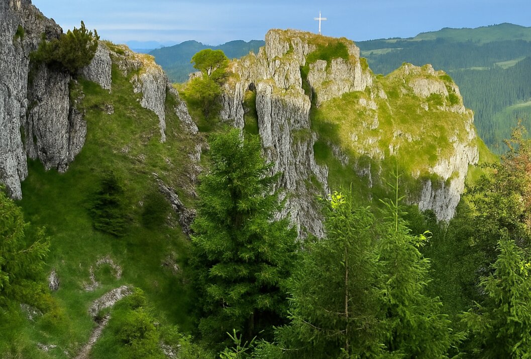 Berglandschaft mit Felsen, Bäumen und einem weißen Kreuz auf dem Gipfel