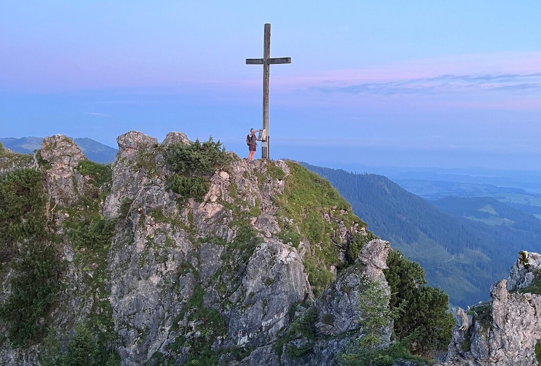 Gipfelkreuz auf felsigem Berg, zwei Personen stehen davor, im Hintergrund Berge und blauer Himmel