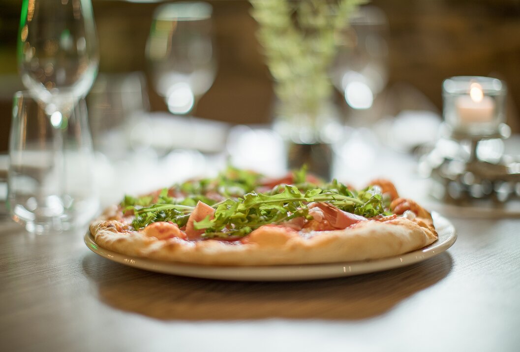 Pizza mit Rucola und Schinken auf Teller, gedeckter Tisch im Hintergrund