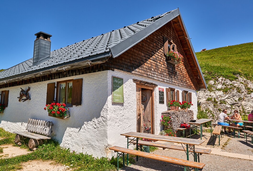 Hütte mit weißen Wänden, braunem Dach und Holzverkleidung, davor Bänke und Tische mit roten Blumen