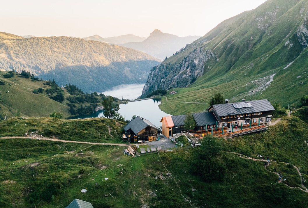 Berghütte am See in grüner Landschaft mit Bergen im Hintergrund