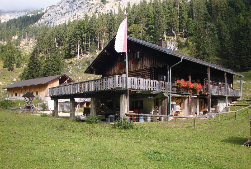 Holzhütte mit Balkon vor Berglandschaft und Wiese, rot-weiße Fahne